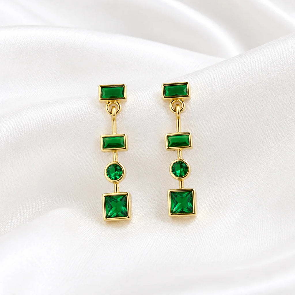 Flat lay of Ma Vie emerald drop earrings on cream silk, 18k gold filled green CZ geometric