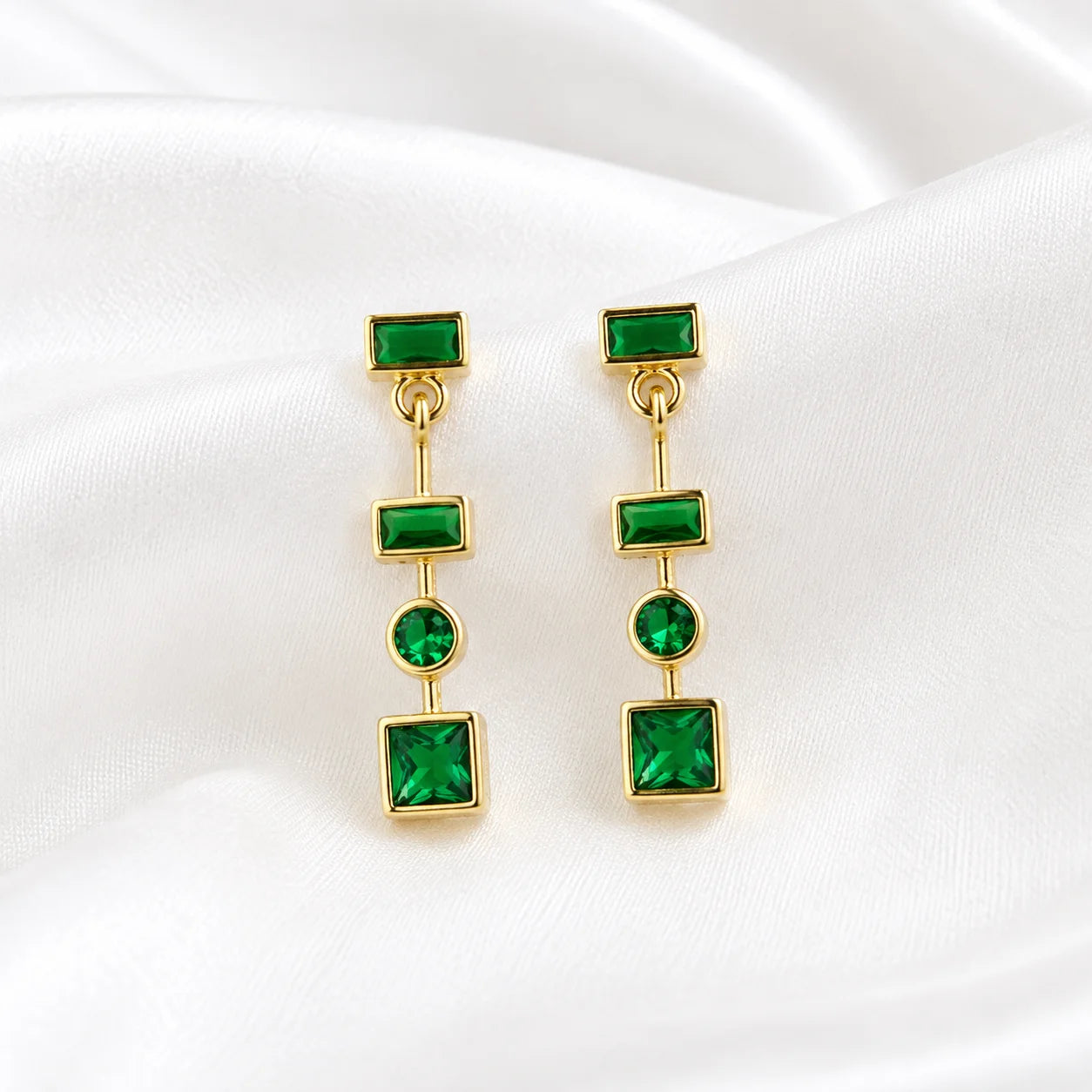 Flat lay of Ma Vie emerald drop earrings on cream silk, 18k gold filled green CZ geometric